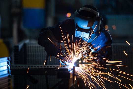 A person wearing protective welding gear is using a welding torch, creating bright sparks while working on a metal structure in a workshop environment.