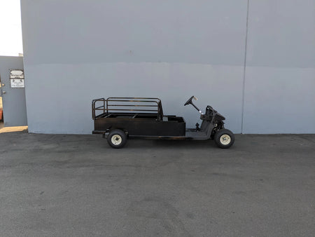 Black utility golf cart with flatbed cargo area and railing parked outside against a plain wall featuring the EZGO-RXV-ST-FLAT-60 EZ GO RXV FLATBED RETROFIT CONVERSION custom fabrication for golf carts.
