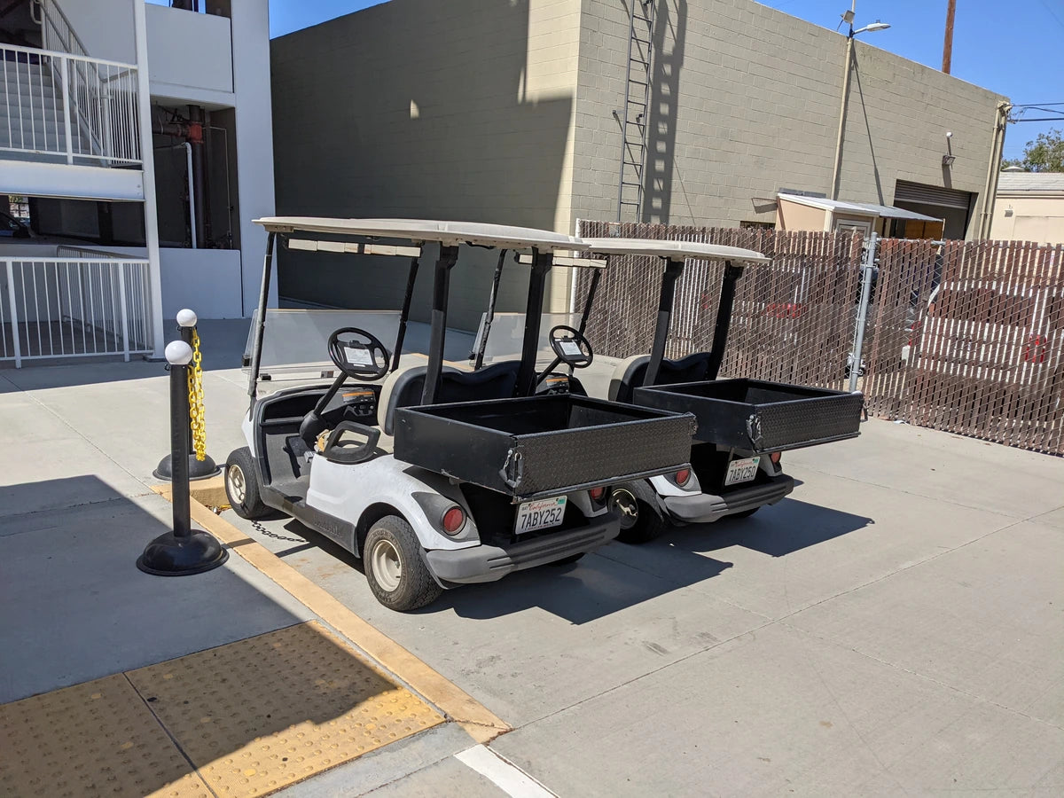 Two golf carts parked side by side, both equipped with YAM-DRIVE-UTIL31 YAMAHA DRIVE HEAVY-DUTY INDUSTRIAL UTILITY BOX WITH TAILGATE; custom fabrication for golf carts.