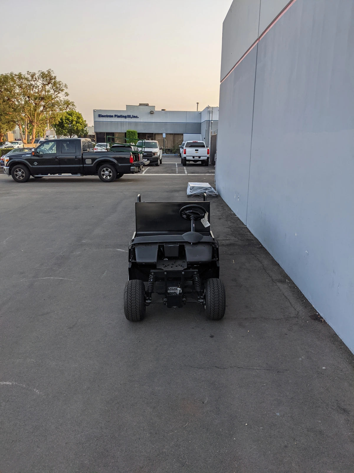 Rear view of a custom black golf cart with a flatbed and convertible seat parked outside near a grey building, showcasing the EZGO-RXV-ST-FLAT-CONV-72 EZ GO RXV FLATBED RETROFIT WITH CONVERTIBLE SEAT designed for golf carts.