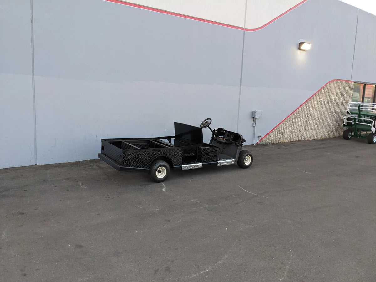 EZGO-RXV-ST-FLAT-CONV-72 EZ GO RXV FLATBED RETROFIT WITH CONVERTIBLE SEAT shown in black parked next to a building, custom fabrication for golf carts featuring a flatbed cargo area and convertible seating.