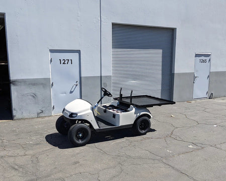 EZGO-TXTVAL-DECK48X48 EZ GO TXT, VALOR, AND MEDALIST DIAMOND PLATE STEEL UTILITY DECK 48″ X 48″ WITH STAKE POCKETS installed on a white golf cart parked outside a warehouse, custom fabrication for golf carts.