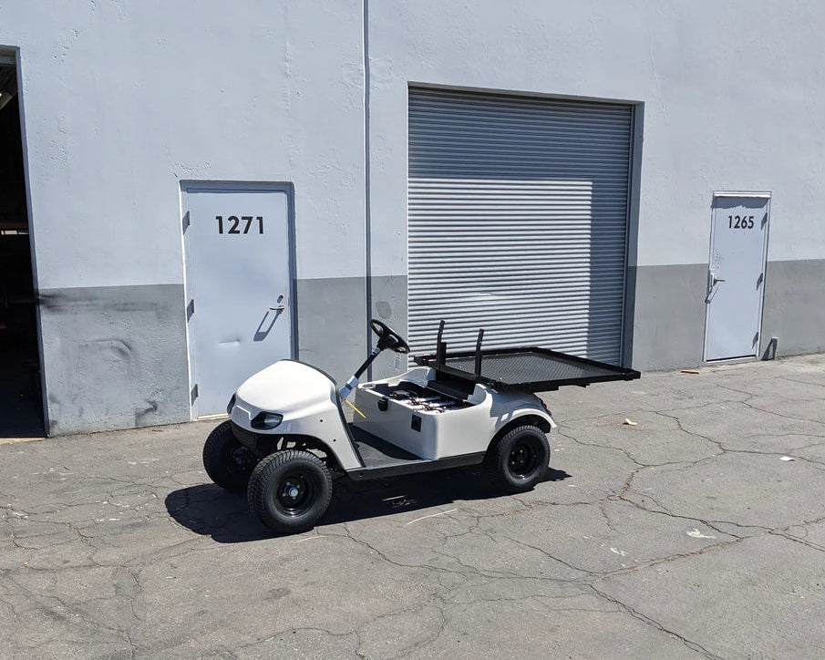 EZGO-TXTVAL-DECK48X48 EZ GO TXT, VALOR, AND MEDALIST DIAMOND PLATE STEEL UTILITY DECK 48″ X 48″ WITH STAKE POCKETS installed on a white golf cart parked outside a warehouse, custom fabrication for golf carts.