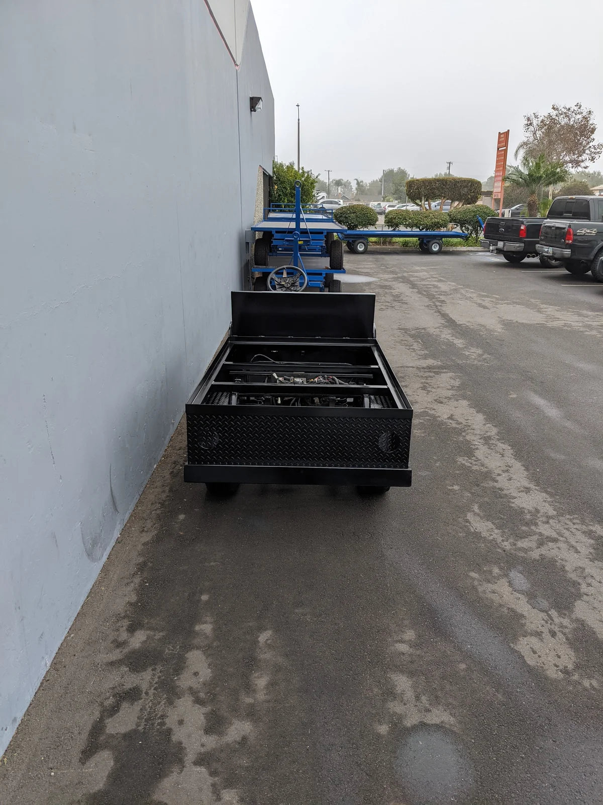 Black metal flatbed utility structure with an open top and storage compartments, parked outside near a blue cart and vehicles, showcasing the YAM-UMAX-ST-FLAT-72 Yamaha Umax 6 foot Flatbed Conversion custom fabrication for golf carts.