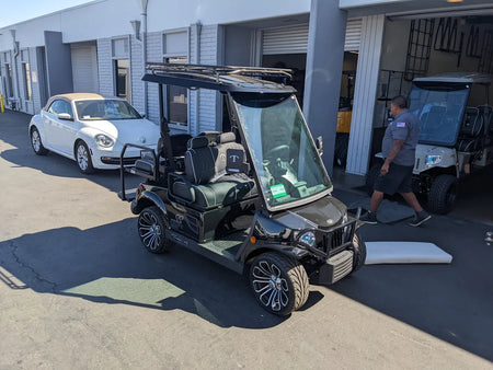 Black custom golf cart equipped with TOM-EMERGE2PASS-49RR3 Tomberlin Emerge 2 Passenger Roof Rack parked outside a garage with a white car and man nearby, showcasing custom fabrication for golf carts.