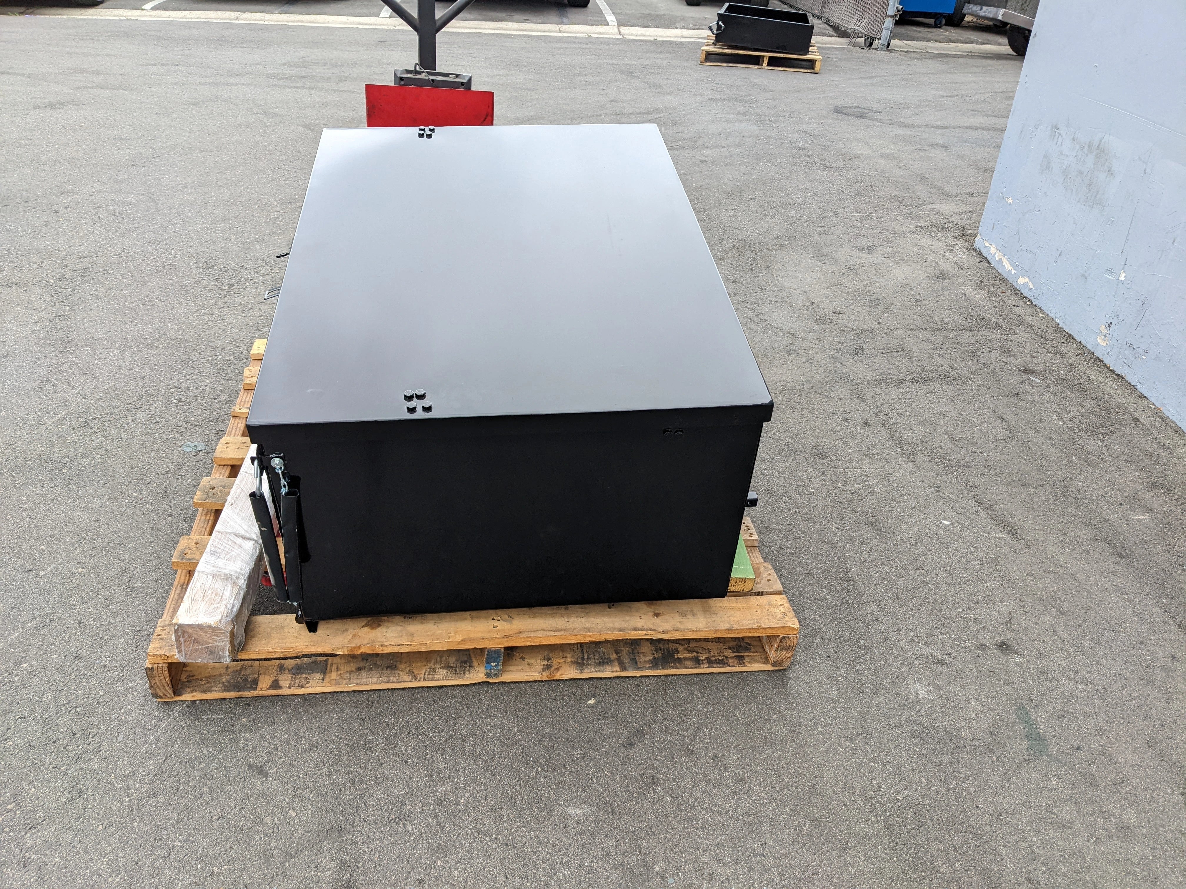Black metal cargo box placed on a wooden pallet outdoors, compatible with CARGO31-GAS-SPRING-RETROFIT GAS SPRING KIT FOR CART HAULERS CARGO BOX LID OPENING AND CLOSING for utility and cargo storage.