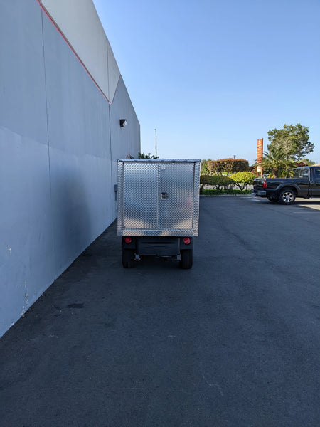Rear view of a custom-fabricated CUSH-HP-CARGO42X48X42-4D CUSHMAN HAULER PRO WITH CAB CARGO BOX WITH LOCKING DOORS mounted on a utility vehicle in an outdoor parking area.