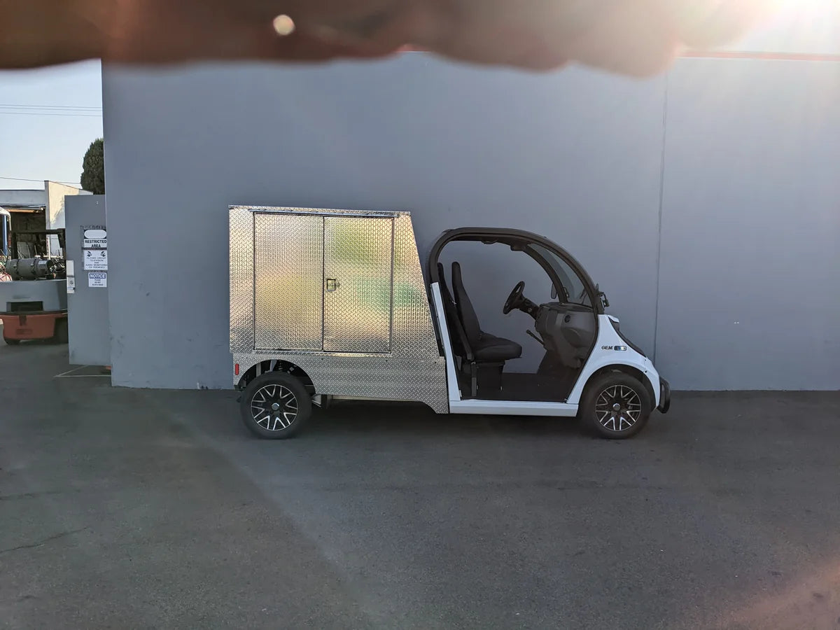White golf cart equipped with a silver metal FMB-GEM-52X70X48-D-D2W0 Multi-Purpose Box for Flatbed, designed for custom fabrication for golf carts, shown outdoors against a gray wall.