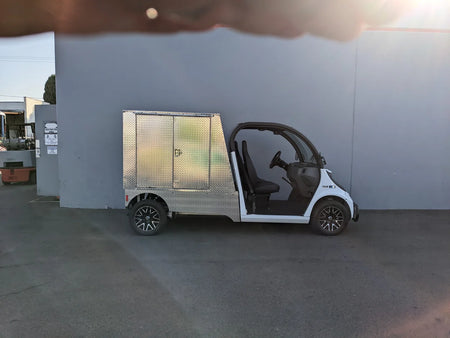 White golf cart equipped with a silver metal FMB-GEM-52X70X48-D-D2W0 Multi-Purpose Box for Flatbed, designed for custom fabrication for golf carts, shown outdoors against a gray wall.