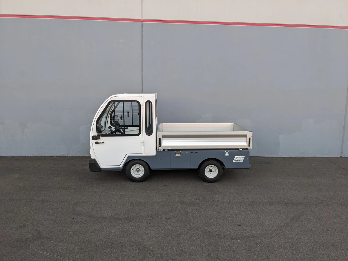 Compact utility vehicle with a flatbed cargo area, shown in profile against a gray wall, featuring the BIGFOOT XL BATTERY SLIDE-OUT RETROFIT FOR TAYLOR-DUNN BIGFOOT XL installation.