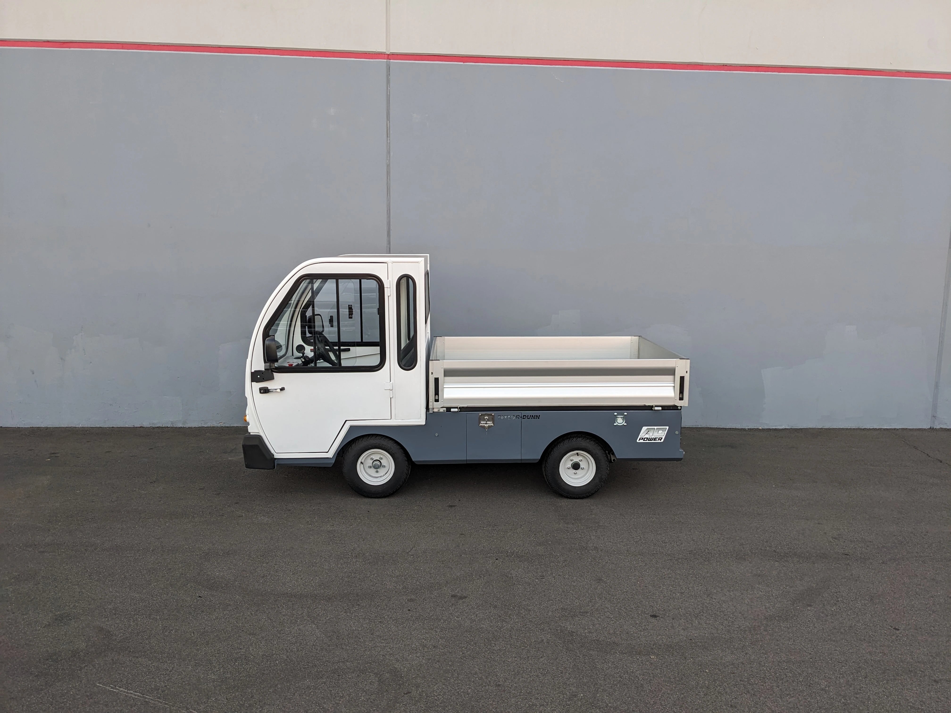 Compact utility vehicle with a flatbed cargo area, shown in profile against a gray wall, featuring the BIGFOOT XL BATTERY SLIDE-OUT RETROFIT FOR TAYLOR-DUNN BIGFOOT XL installation.