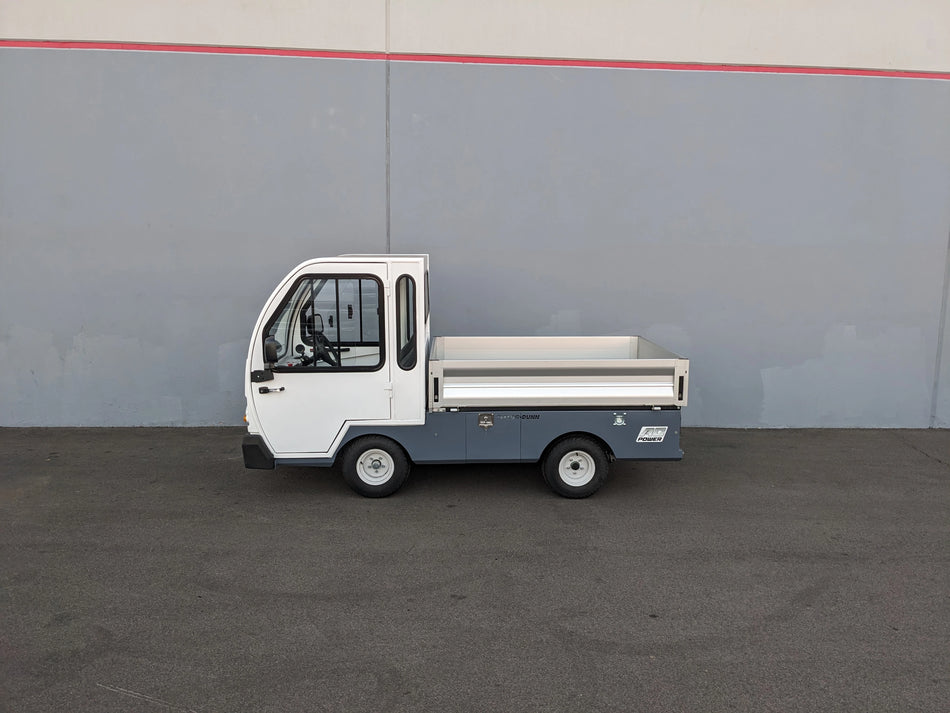 Compact utility vehicle with a flatbed cargo area, shown in profile against a gray wall, featuring the BIGFOOT XL BATTERY SLIDE-OUT RETROFIT FOR TAYLOR-DUNN BIGFOOT XL installation.