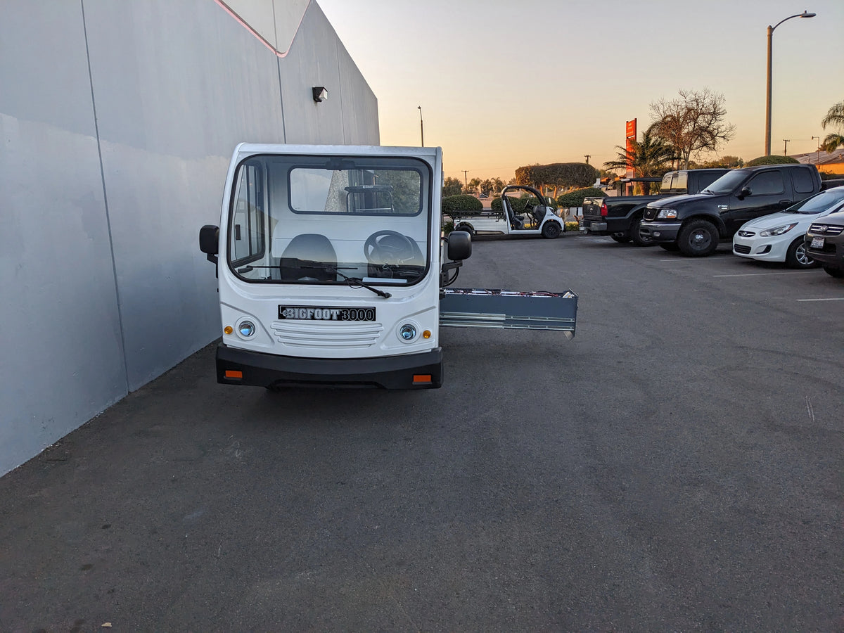 White utility vehicle with a slide-out platform extended, illustrating the BIGFOOT XL BATTERY SLIDE-OUT RETROFIT FOR TAYLOR-DUNN BIGFOOT XL parked outside near other vehicles.