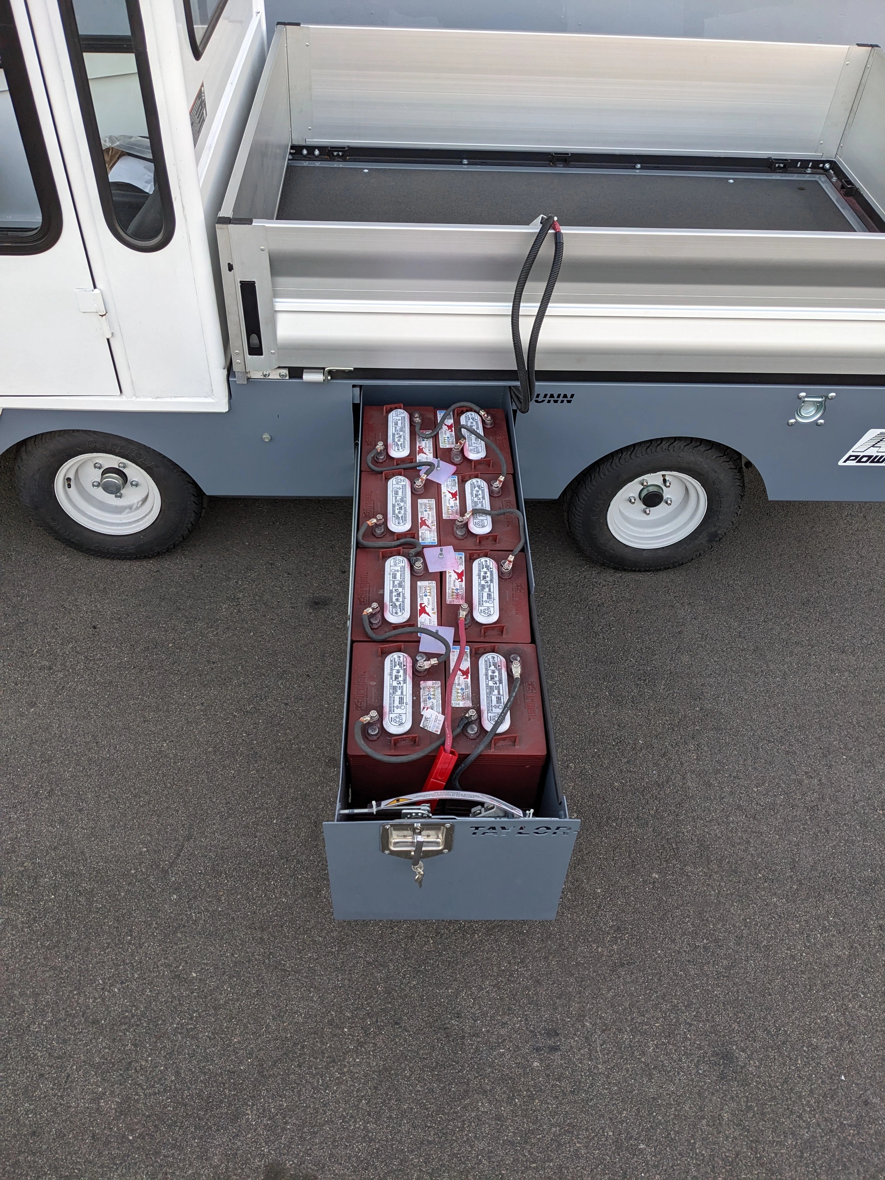 BIGFOOT XL BATTERY SLIDE-OUT RETROFIT FOR TAYLOR-DUNN BIGFOOT XL showing a metal battery slide-out tray with multiple connected batteries installed on a utility vehicle with a flatbed cargo area.