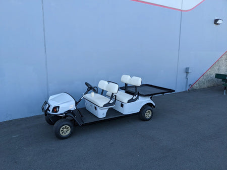 White golf cart equipped with CUSH-SHUTTLE-DECK48X48-SP CUSHMAN SHUTTLE CARGO DECK WITH STAKE POCKETS flatbed cargo area, custom fabrication for golf carts, shown parked against a blue wall.