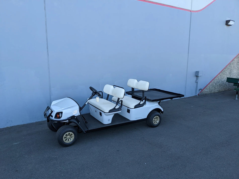 White golf cart equipped with CUSH-SHUTTLE-DECK48X48-SP CUSHMAN SHUTTLE CARGO DECK WITH STAKE POCKETS flatbed cargo area, custom fabrication for golf carts, shown parked against a blue wall.