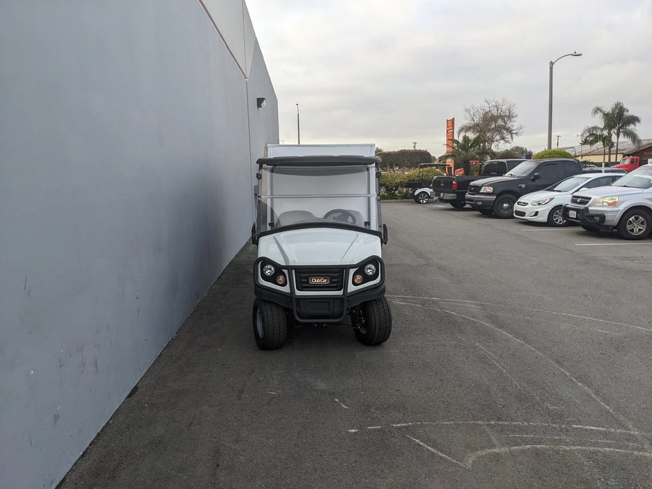 Small white utility golf cart parked beside a gray building with other vehicles in the background, associated with CC-CARRYALL900-LAUNDRY BOX FLATBED SPECIFIC PURPOSE BOX FOR CARRYALL 900 WITH LOCKING DOORS, SHELVES, AND HANGING ROD.
