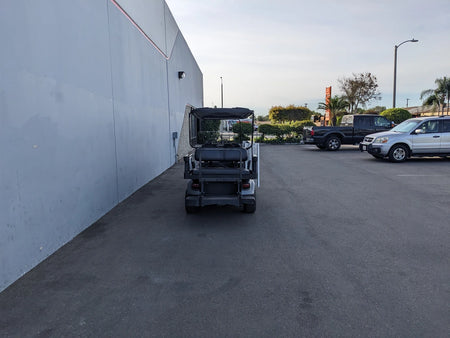 Rear view of a golf cart customized with CUSH-WHEELCHAIR-TRANSPORT CUSHMAN GOLF CART CONVERSIONS FOR WHEELCHAIR PASSENGER, parked near a building with other vehicles nearby, showcasing custom fabrication for golf carts.