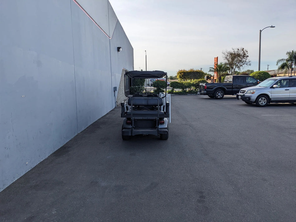 Rear view of a golf cart customized with CUSH-WHEELCHAIR-TRANSPORT CUSHMAN GOLF CART CONVERSIONS FOR WHEELCHAIR PASSENGER, parked near a building with other vehicles nearby, showcasing custom fabrication for golf carts.