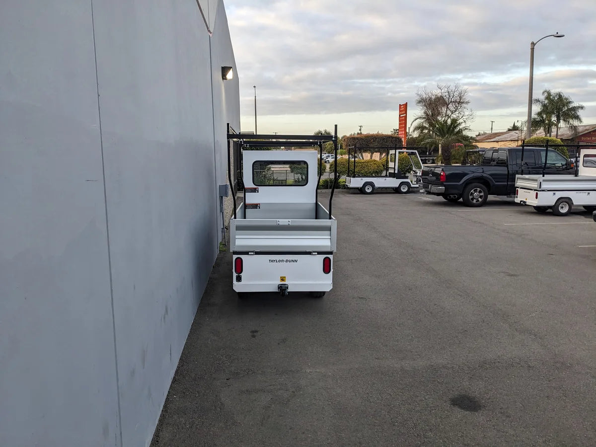 White utility golf cart equipped with TD-BIG-44X120-TLRI Top Mount Ladder Rack for mounting inside the bed of a Taylor Dunn Bigfoot, custom fabrication for golf carts in a parking lot.