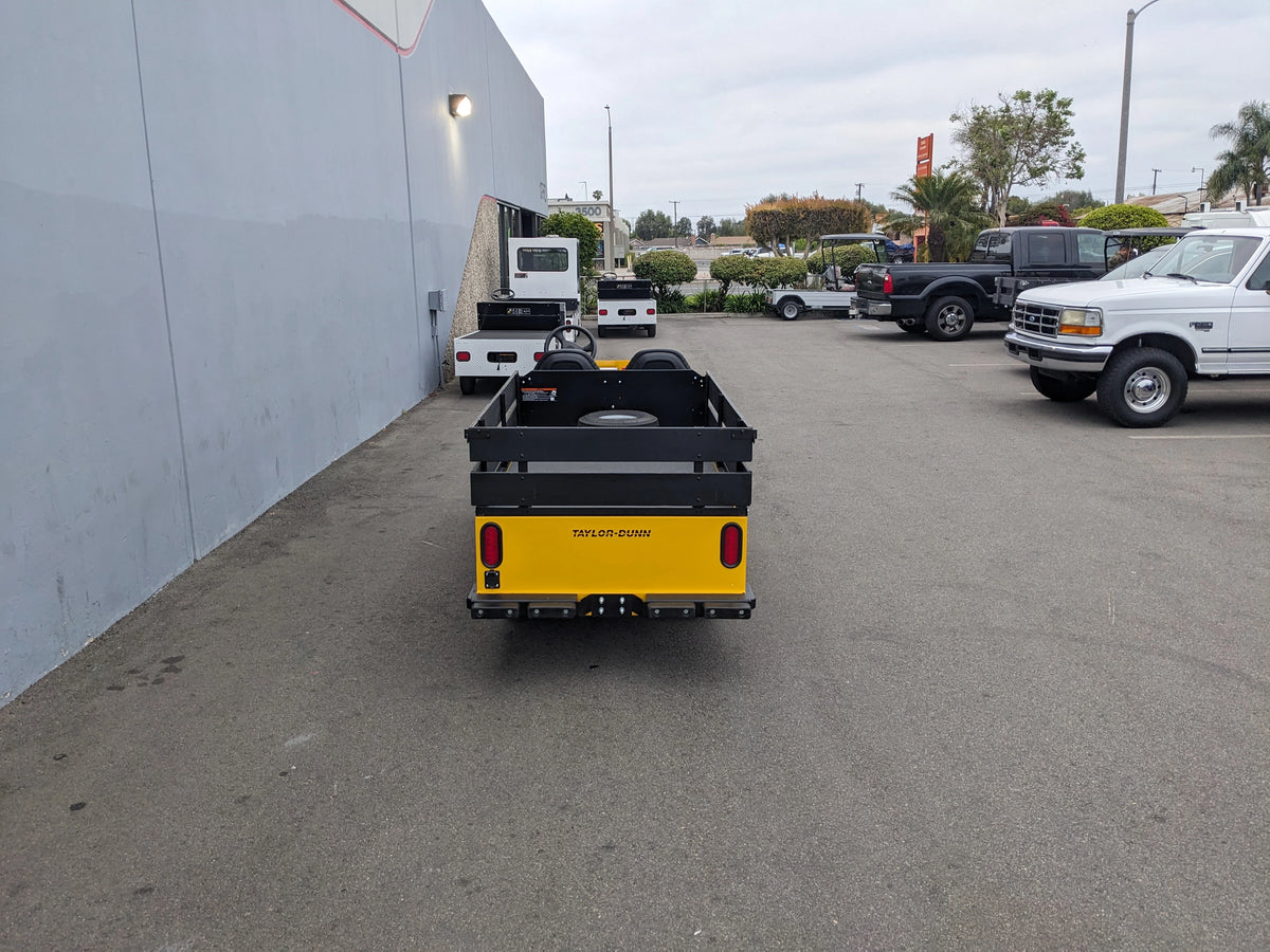 Rear view of a utility vehicle equipped with the BIGFOOT-DOOR-COMPARTMENT Pass Through Storage Cabinet for Taylor Dunn Bigfoot, parked in an outdoor lot alongside trucks and industrial equipment.