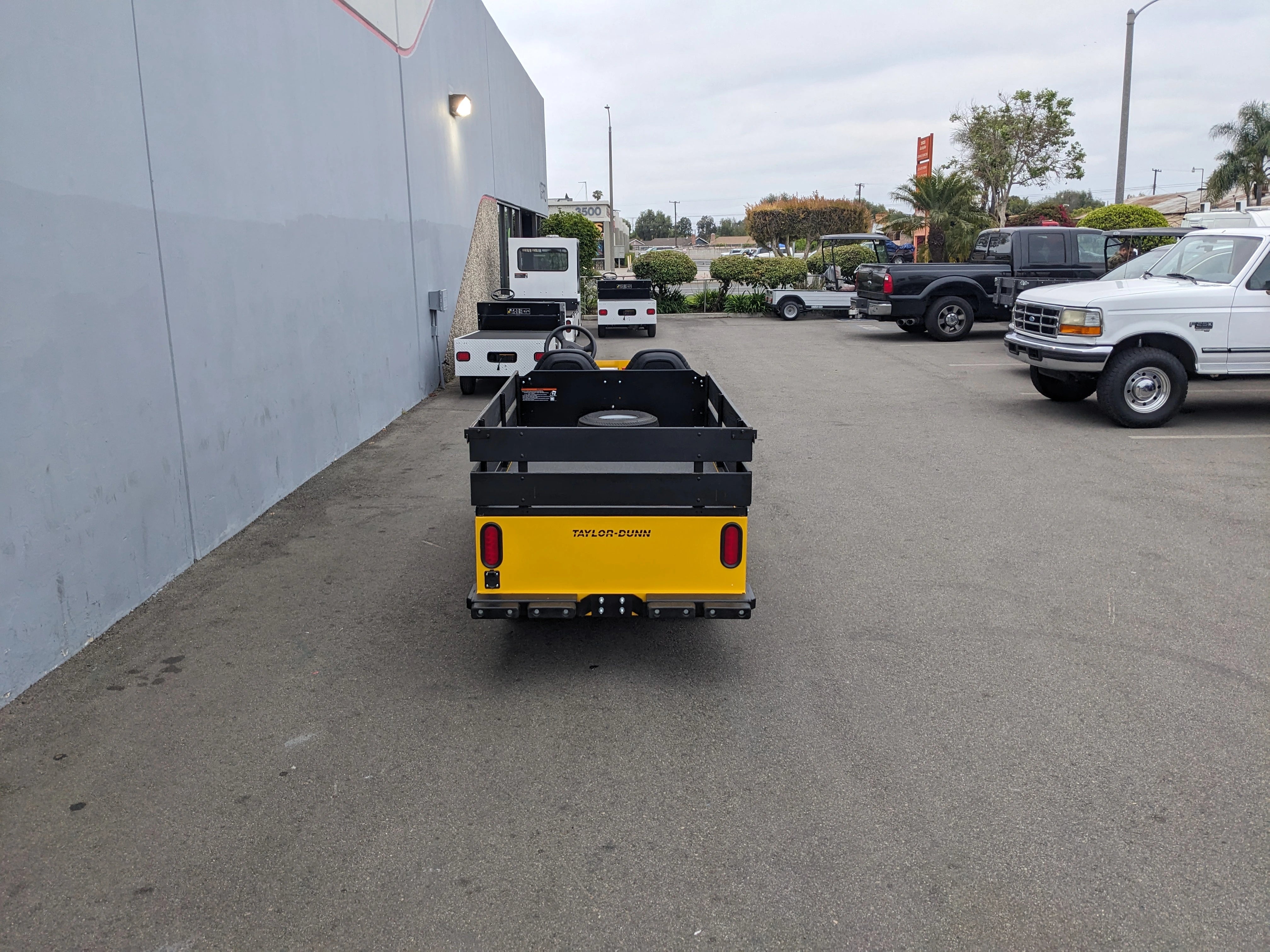 Rear view of a utility vehicle equipped with the BIGFOOT-DOOR-COMPARTMENT Pass Through Storage Cabinet for Taylor Dunn Bigfoot, parked in an outdoor lot alongside trucks and industrial equipment.