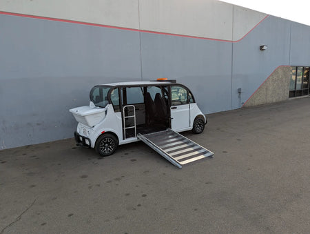 White golf cart with an extended metal ramp parked beside a building, custom fabricated for wheelchair access as a GEM-E6 WHEELCHAIR TRANSPORT GEM GOLF CART CONVERSION FOR WHEELCHAIR PASSENGER.