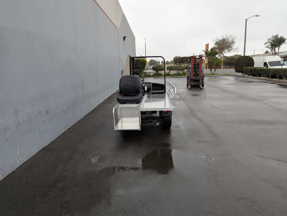 Rear view of a custom fabricated CUSH-EMT-84X24 EMT GOLF CART BED CONVERSION FOR CUSHMAN GOLF CART showing a flatbed with metal storage boxes and a black seat, parked outdoors on a wet pavement near industrial buildings.