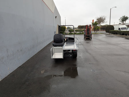 Rear view of a custom fabricated CUSH-EMT-84X24 EMT GOLF CART BED CONVERSION FOR CUSHMAN GOLF CART showing a flatbed with metal storage boxes and a black seat, parked outdoors on a wet pavement near industrial buildings.