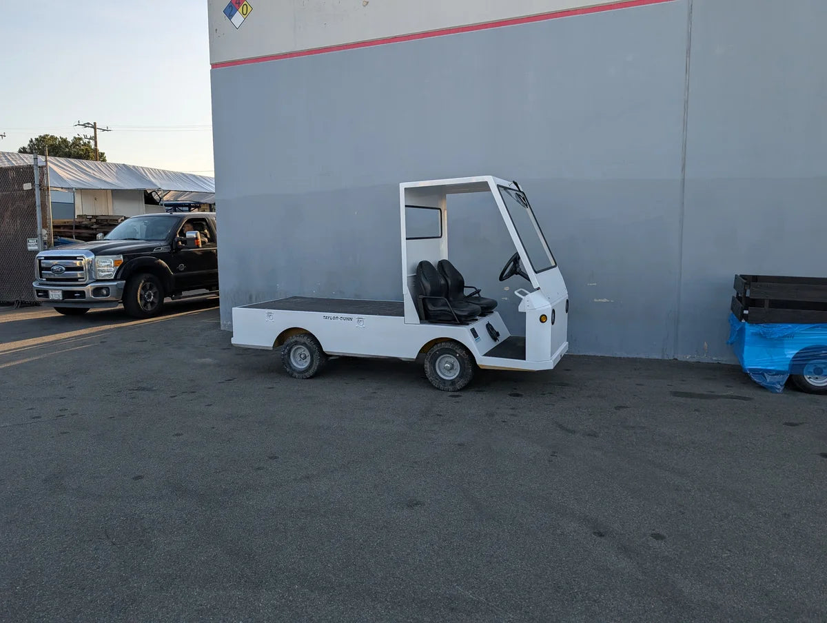 SCE-TDBIG-CAB-ND CAB ENCLOSURE FOR TAYLOR DUNN BIGFOOT, NO DOORS electric utility vehicle with a flatbed, designed for custom fabrication for golf carts, parked outdoors by a building.